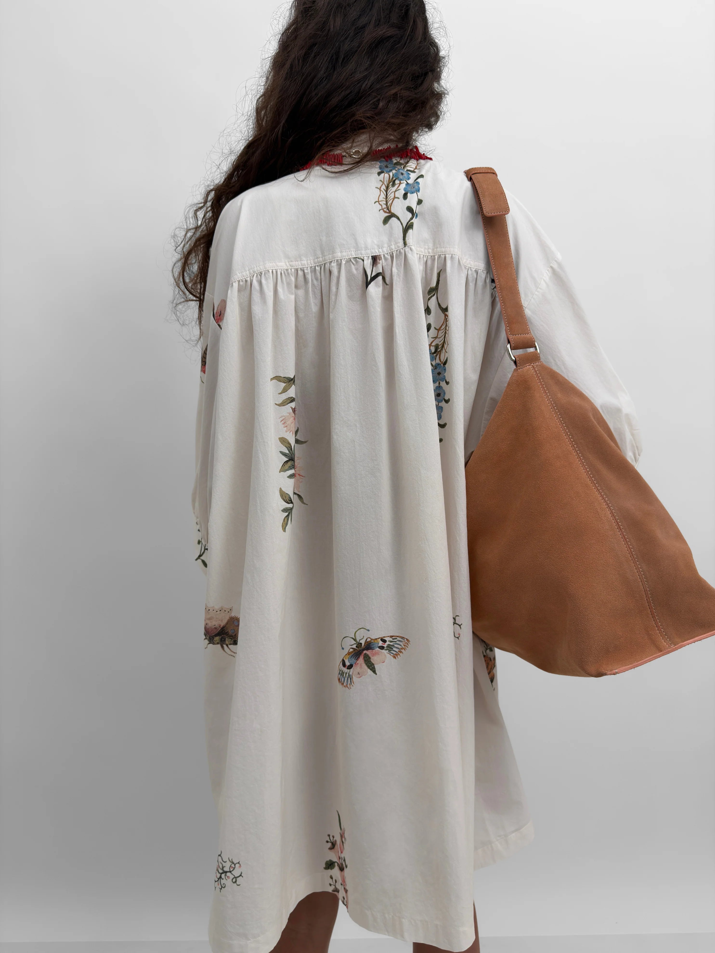 Woman wearing a cream-colored dress with floral print, holding a brown bag against a white background.