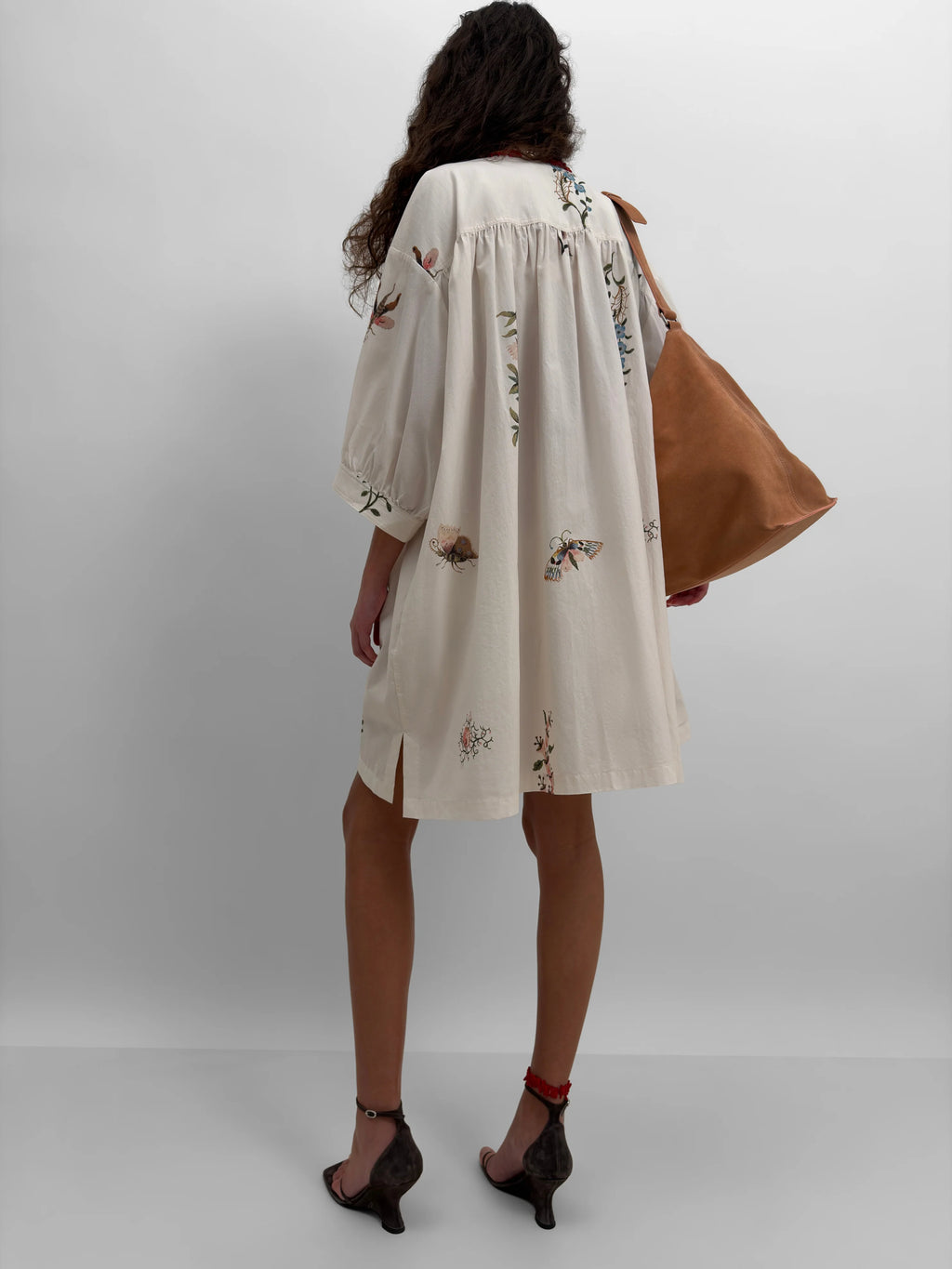Woman wearing a cream dress with floral print and holding a brown bag on a white background