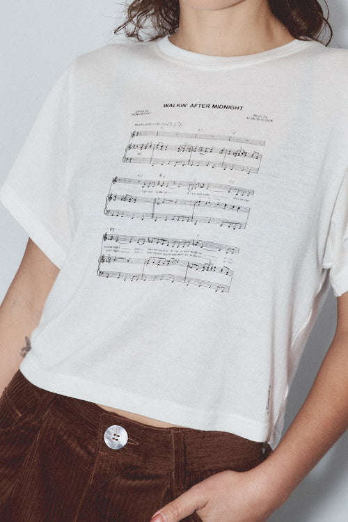 A woman wearing a white short-sleeve t-shirt with black musical notation text printed on the front, by Fashion Brand Company paired with dark brown corduroy pants, standing against a light gray background.