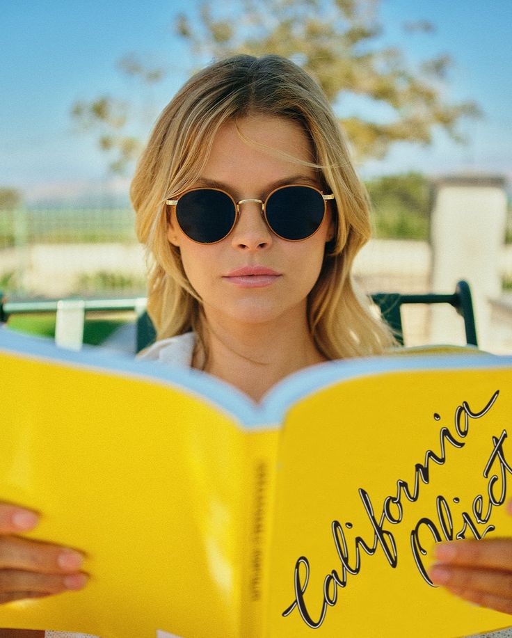 Woman wearing sunglasses and reading a yellow book titled 'California' outdoors.