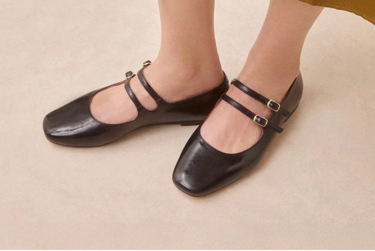 Black flat shoes with strap worn by a person in a mustard yellow skirt on a beige background