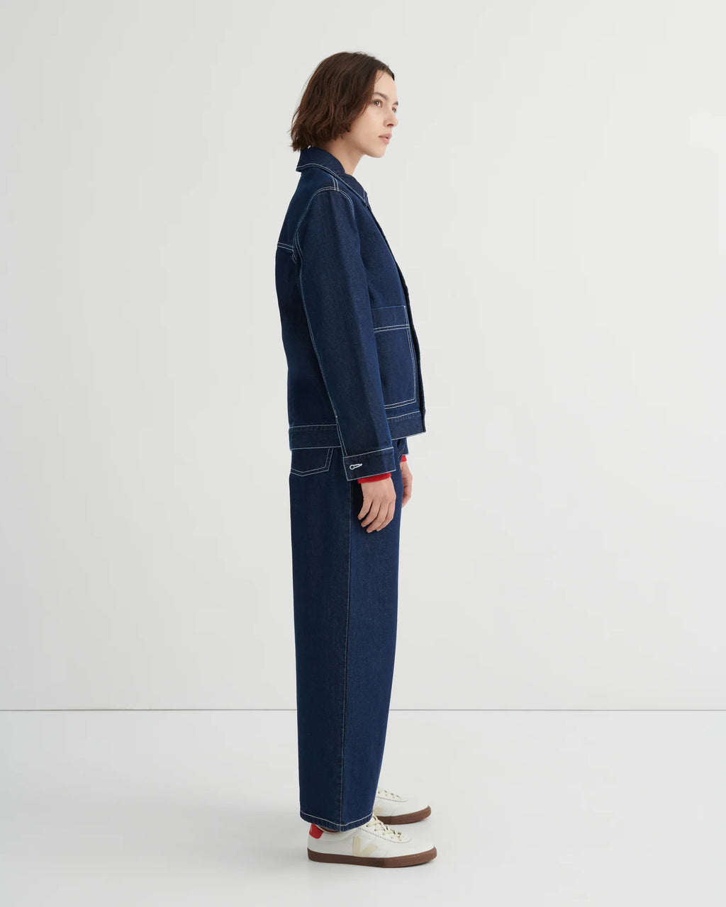 A model shown in profile wearing a dark blue denim Worker jacket by Kowtow and matching wide-leg denim pants, paired with white sneakers, against a clean white studio background.