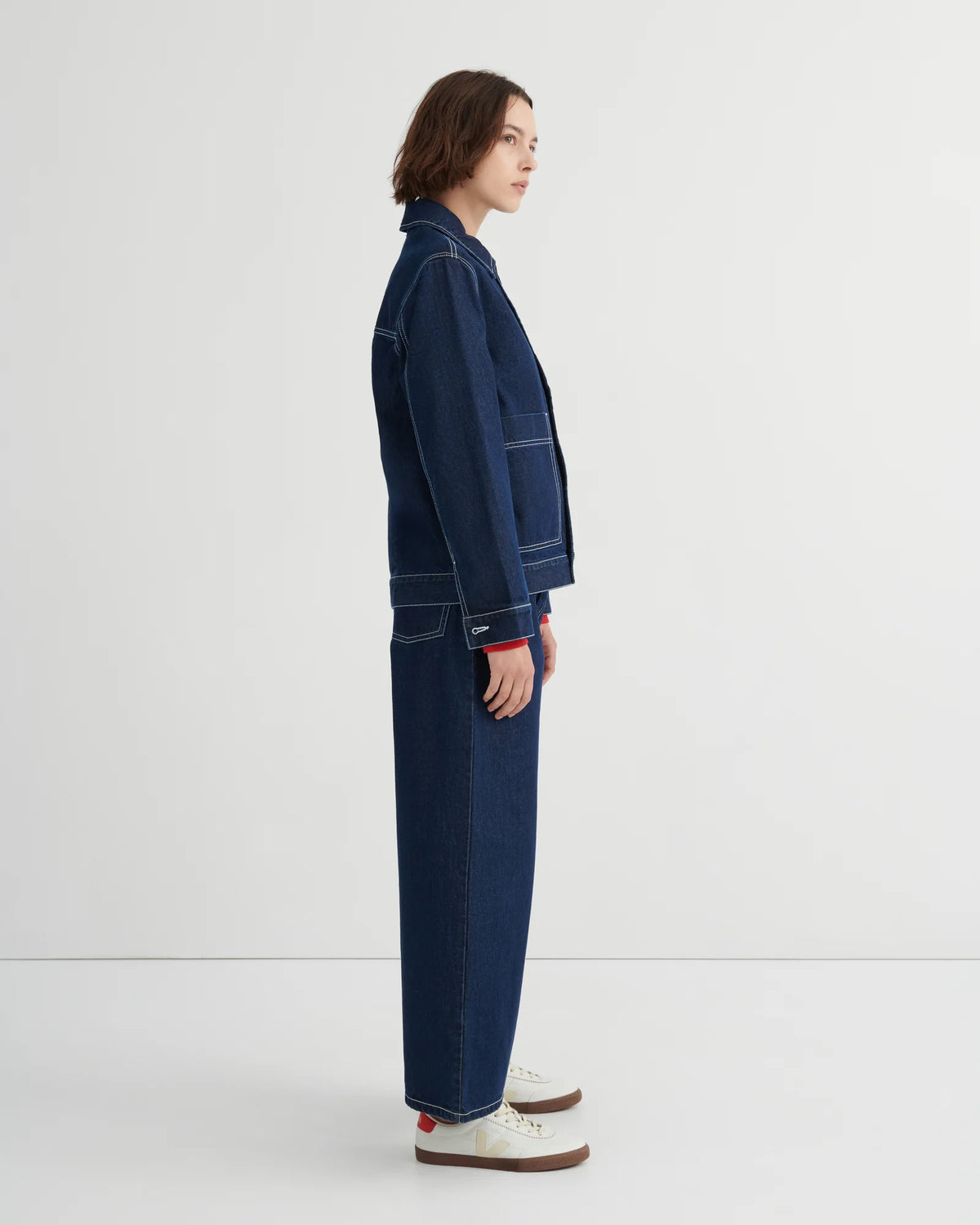 A model shown in profile wearing a dark blue denim Worker jacket by Kowtow and matching wide-leg denim pants, paired with white sneakers, against a clean white studio background.