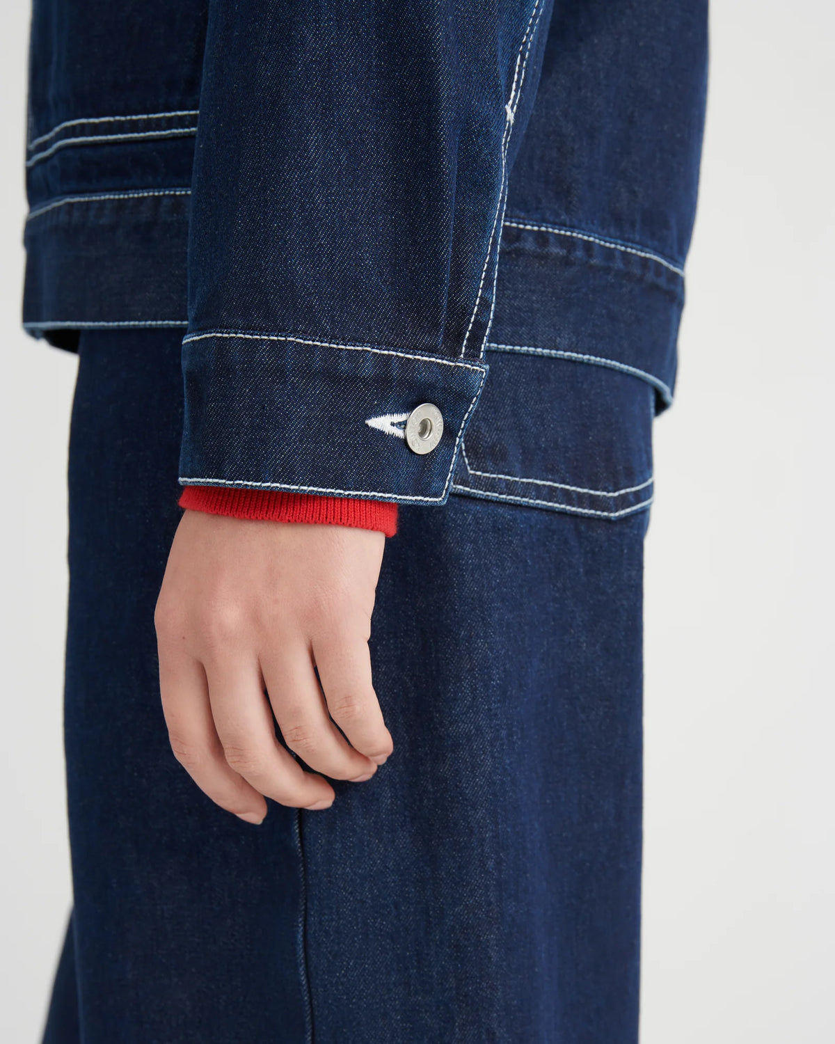 Close-up of a person wearing a dark indigo denim jacket and matching denim skirt or pants, showing the sleeve cuff with white contrast stitching, a silver button, and a red ribbed knit undersleeve.