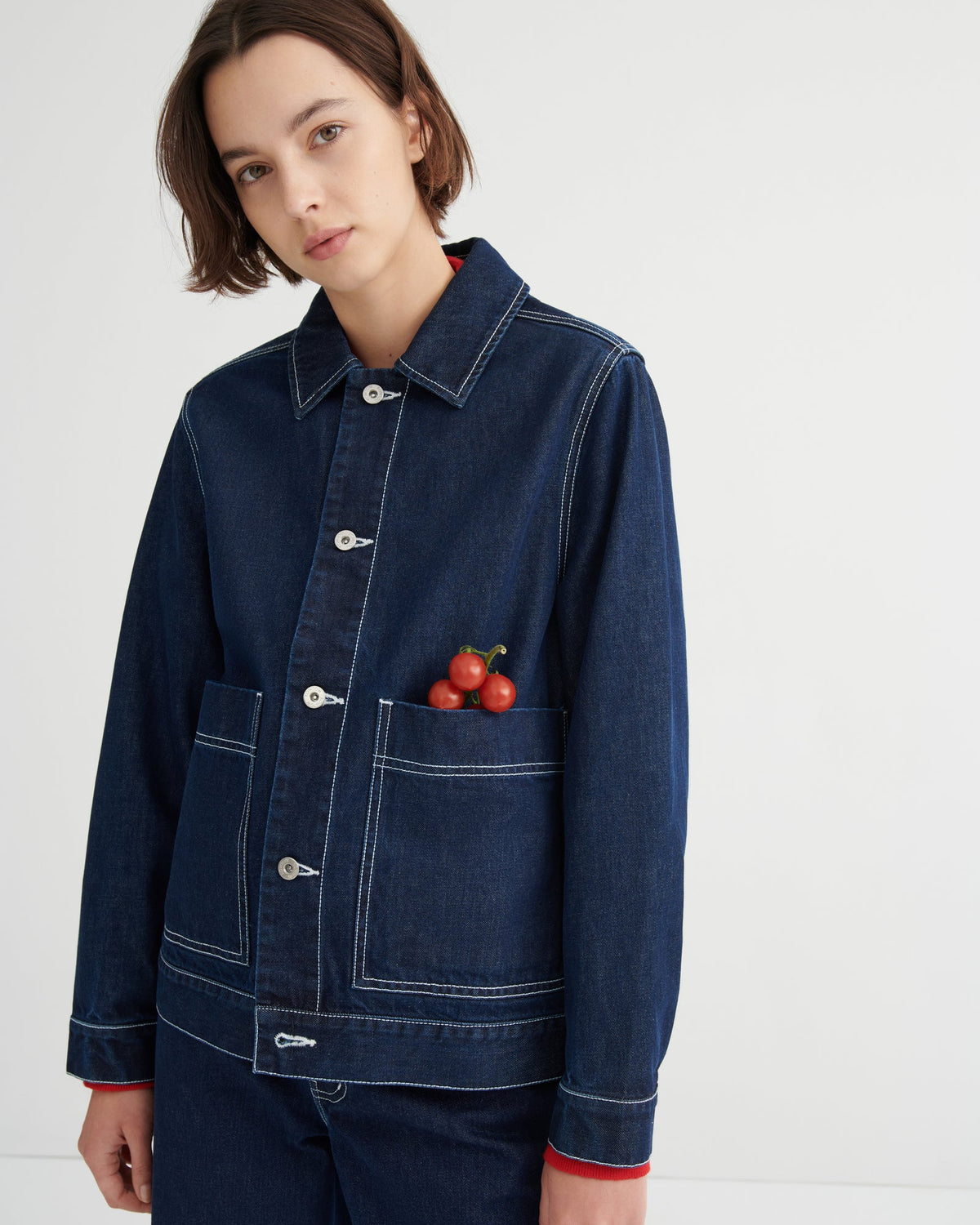 A model wearing a dark blue denim jacket Worker Jacket by Kowtow with white contrast stitching, silver snap buttons, and a front pocket holding cherry tomatoes, paired with matching dark denim pants against a plain white studio background.